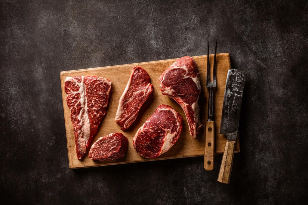 Several large cuts of steak rest of a cutting board along with prongs and a large knife